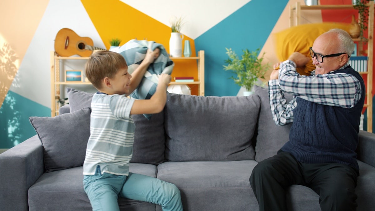 Grandfather old man is playing with joyful kid fighting pillows having fun in apartment enjoying game together. Lifestyle, people and happy family concept.